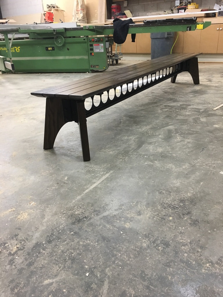 Long wooden bench with circular cutouts on a concrete floor in an industrial setting.